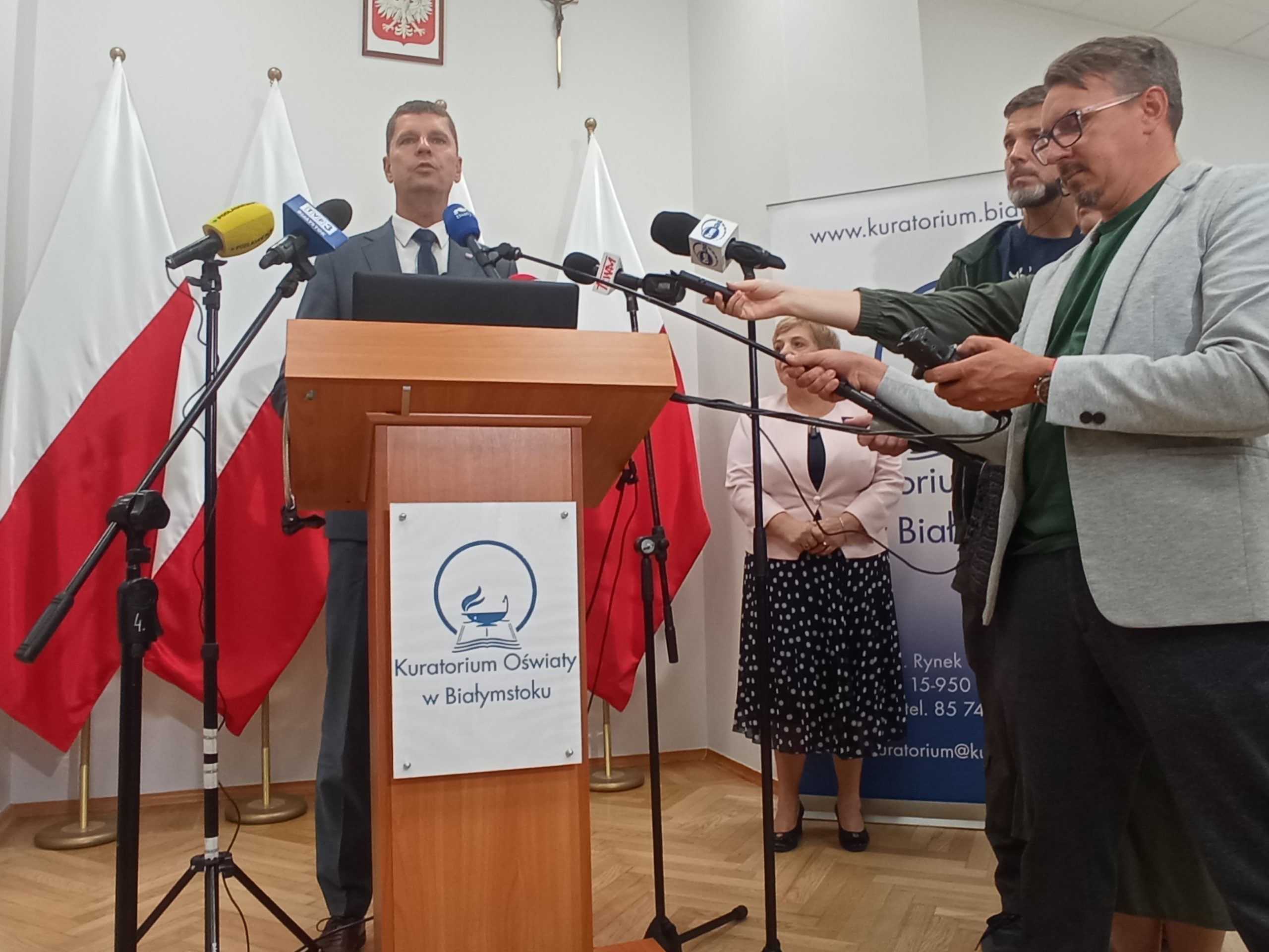 Mężczyzna stoi przy mikrofonach, obok stoją dziennikarze