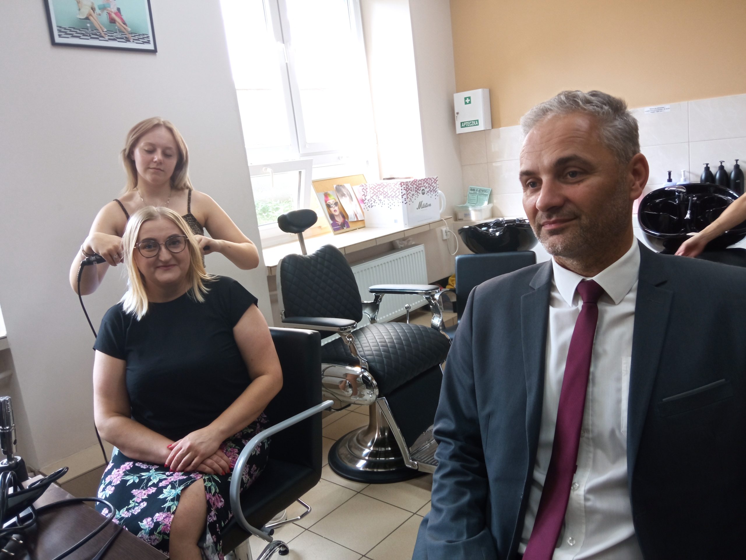 NA fryzjerskich krzesłach siedzą kobieta i mężczyzna, młoda dziewczyna układa włosy kobiety