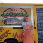 Mężczyzna w kuchennym fartuchu stoi przed food truckiem