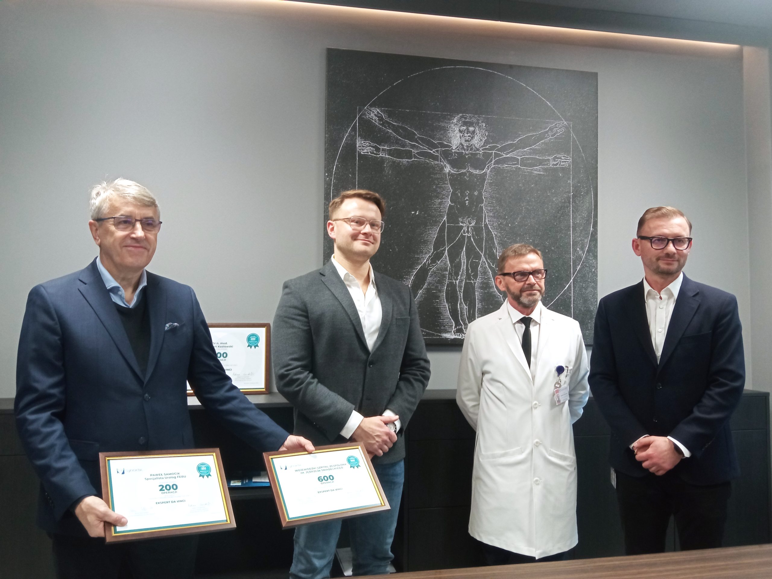 Czterech mężczyzn stoi na tle grafiki Leonarda Da Vinci, jeden trzyma dyplomy