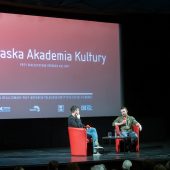 Telebim na scenie kinowej, na której siedzą mężczyźni.