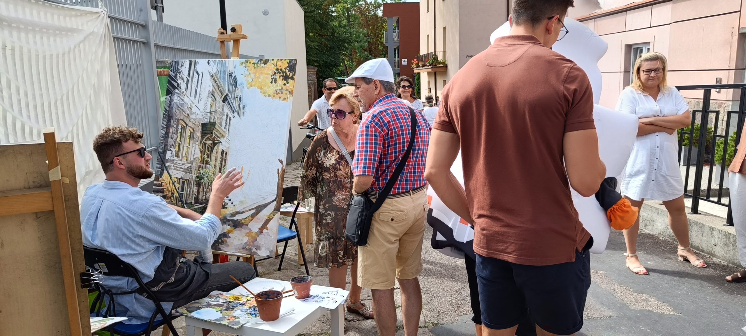 kilka osób stoi na chodniku, po lewej stronie na sztaludze znajduje się obraz