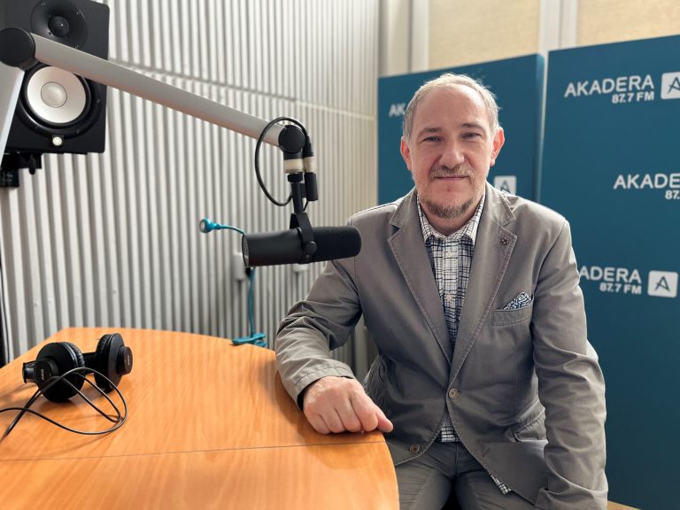 starszy mężczyzna z siwym zarostem siedzi przy stole z mikrofonem, ma szarą marynarkę i koszulę w kratę