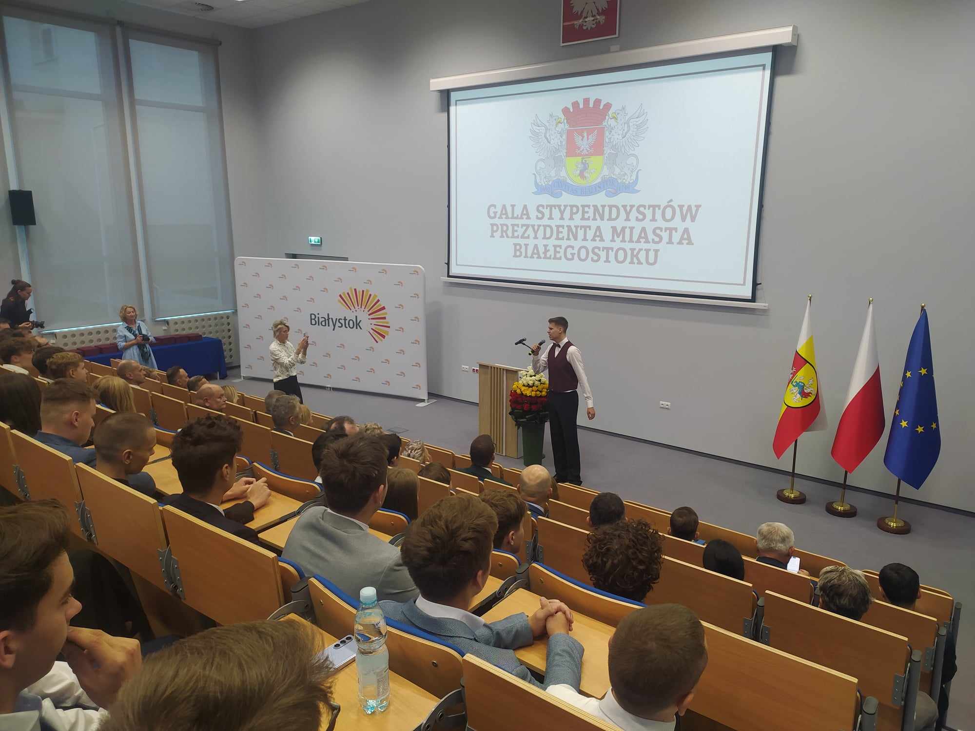 Na auli siedzą ludzie, a przed nim mężczyzna śpiewa do mikrofonu.