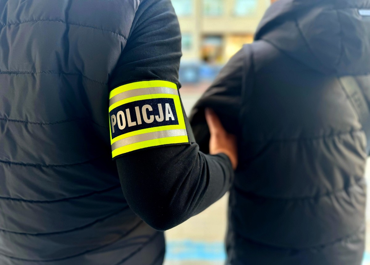 dwie osoby w kurtkach stoją tyłem, jedna na ramieniu ma opaskę z napisem Policja