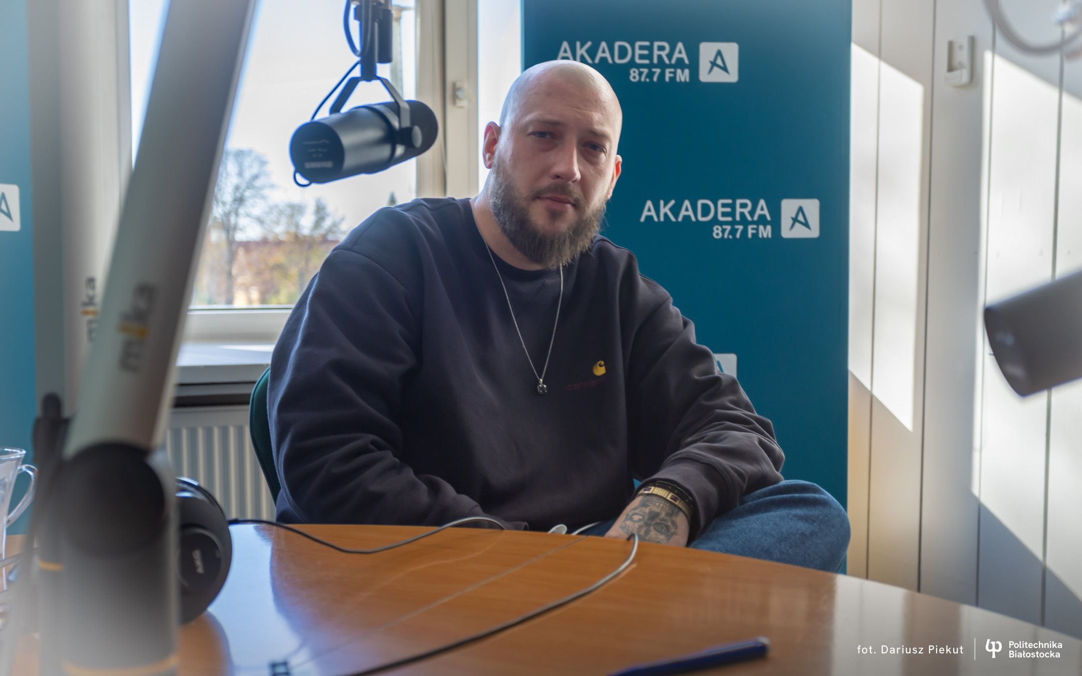 Mężczyzna z brodą i w ciemnej bluzie bez włosów na głowie siedzi za mikrofonem