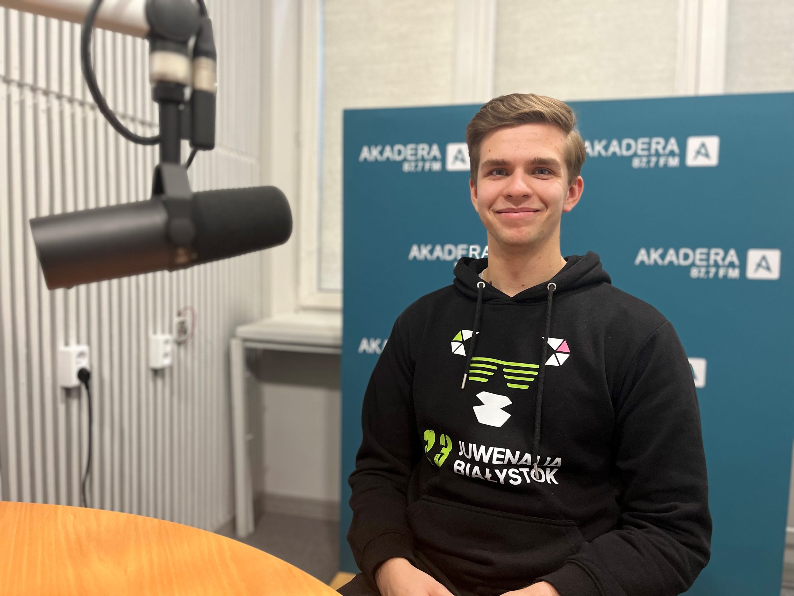 młody student siedzi w czarnej bluzie, uśmiechnięty, włosy ciemne zaczesane na bok