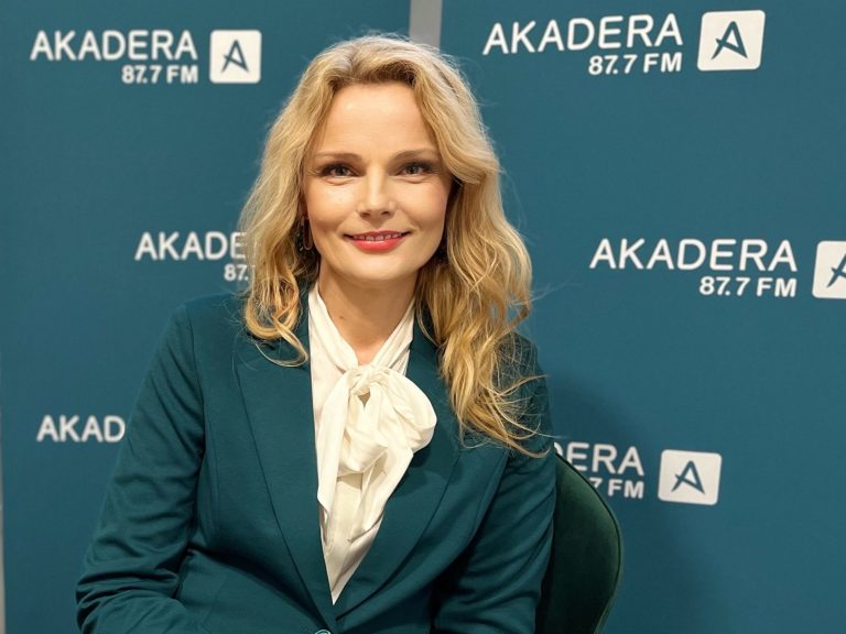 Prof. Katarzyna Halicka podczas wizyty w Radiu AKADERA, siedzi na tle niebieskich plansz z logo stacji, uśmiechnięta, w zielonej marynarce i białej bluzce z kokardą