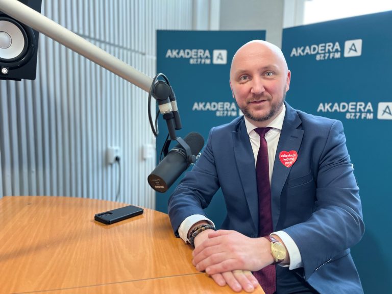 młody mężczyzna z lekkim zarostem, w garniturze i pod krawatem siedzi przy mikrofonie