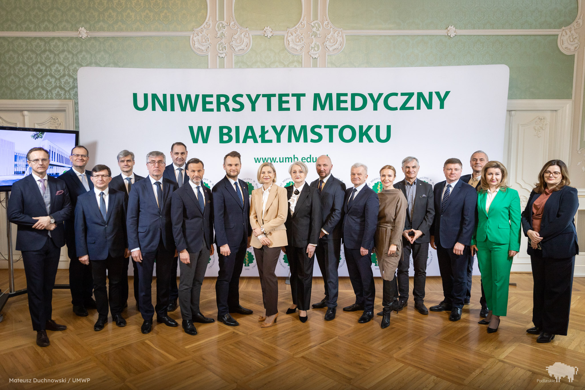 Grupa kilkunastu osób w eleganckich strojach stoi na tle dużego baneru z napisem "Uniwersytet Medyczny w Białymstoku". W tle widoczna jest klasyczna, zdobiona sala konferencyjna. Wśród uczestników znajdują się przedstawiciele władz województwa, parlamentarzyści, rektorzy oraz dyrektorzy szpitali.