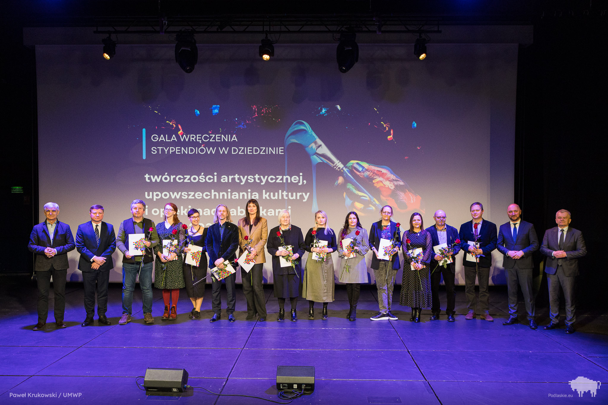 Grupa osób stojących na scenie podczas gali wręczenia stypendiów w dziedzinie twórczości artystycznej, upowszechniania kultury oraz opieki nad zabytkami. W tle duży ekran z grafiką przedstawiającą pędzel malarski oraz tekst informujący o wydarzeniu. Część laureatów trzyma w rękach dyplomy i kwiaty.