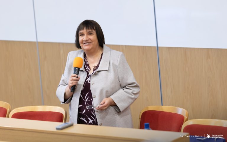 Na zdjęciu znajduje się kobieta w średnim wieku, stojąca w sali wykładowej. Ma krótkie ciemne włosy i uśmiech na twarzy. Ubrana jest w jasną marynarkę i ciemną bluzkę z białym wzorem. Trzyma mikrofon z pomarańczową osłoną i okulary w drugiej ręce. W tle widać drewnianą ścianę i rząd czerwonych krzeseł. Na dole zdjęcia znajduje się podpis fotografa oraz logo Politechniki Białostockiej.