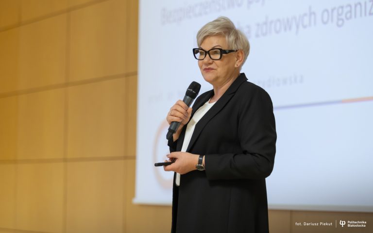 Na zdjęciu prof. dr hab. Joanna M. Moczydłowska z Wydziału Inżynierii Zarządzania Politechniki Białostockiej wygłasza wykład na temat bezpieczeństwa psychologicznego pracowników jako cechy zdrowych organizacji. Profesor stoi przed publicznością, trzyma mikrofon i pilot do prezentacji, a za nią widoczny jest ekran z tytułem wystąpienia. Fotografia wykonana w trakcie wydarzenia na Politechnice Białostockiej.