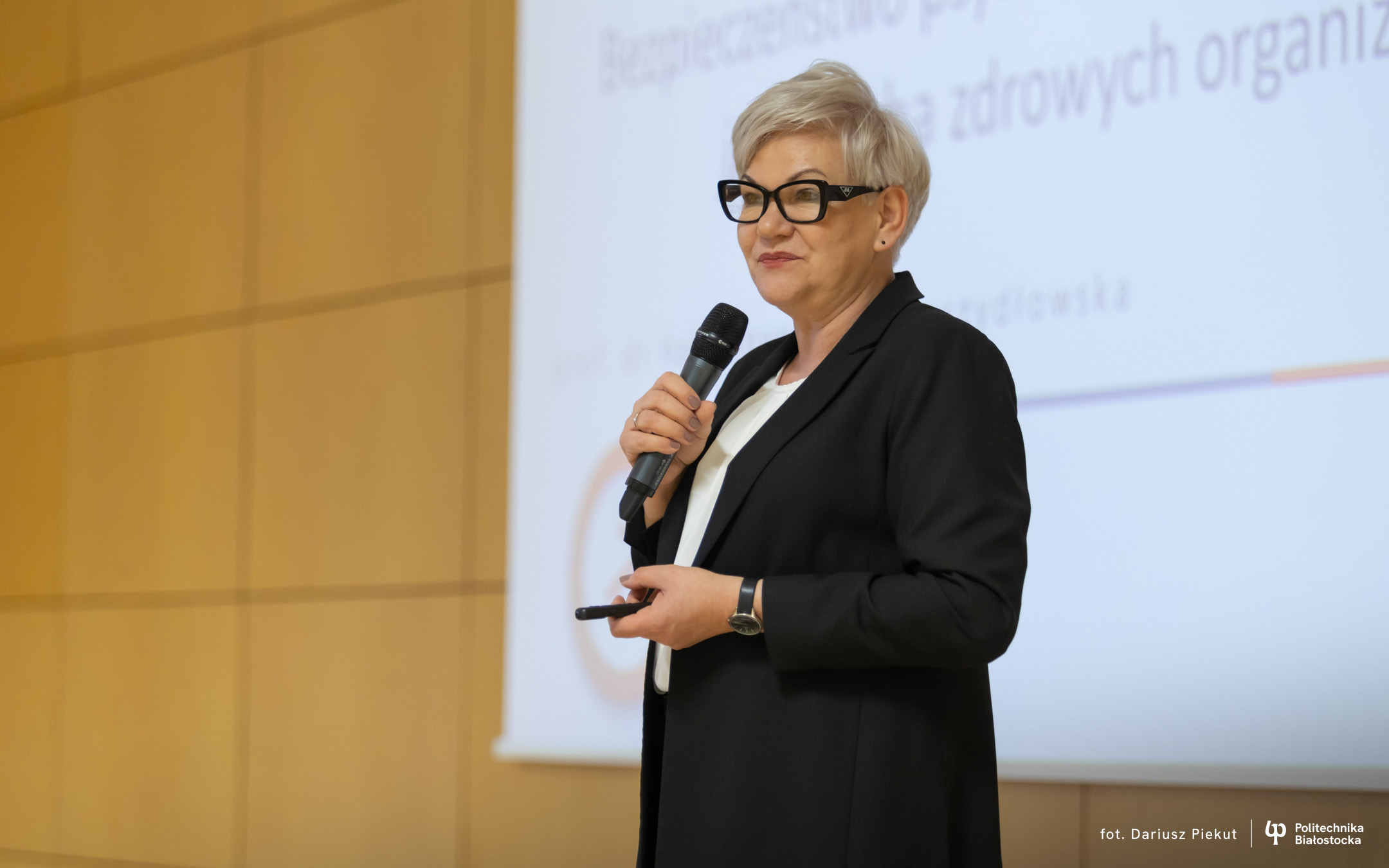Na zdjęciu prof. dr hab. Joanna M. Moczydłowska z Wydziału Inżynierii Zarządzania Politechniki Białostockiej wygłasza wykład na temat bezpieczeństwa psychologicznego pracowników jako cechy zdrowych organizacji. Profesor stoi przed publicznością, trzyma mikrofon i pilot do prezentacji, a za nią widoczny jest ekran z tytułem wystąpienia. Fotografia wykonana w trakcie wydarzenia na Politechnice Białostockiej.