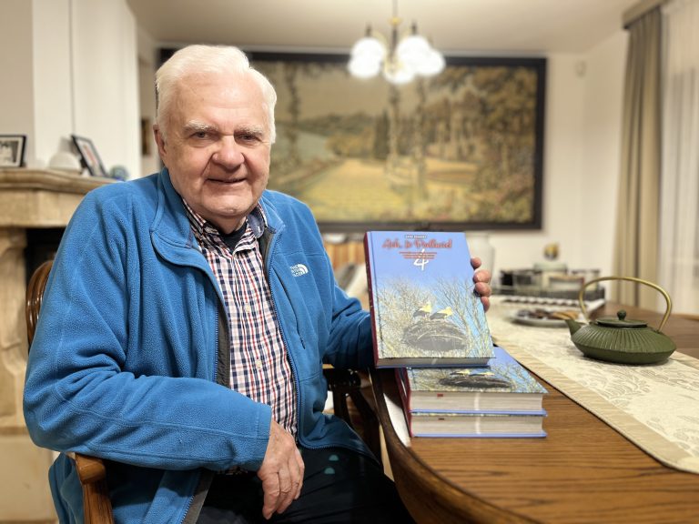 Uśmiechnięty Lech Pilecki siedzi przy stole w swoim domu, trzymając w ręku najnowszy – czwarty – tom książki Ach, to Podlasie! Podróże do niebanalnych i niesamowitych ludzi, miejsc i budowli regionu. Przed nim leży stos wcześniejszych tomów serii. W tle widać stylowy kominek, stół z imbrykiem i gobelin z pejzażem.