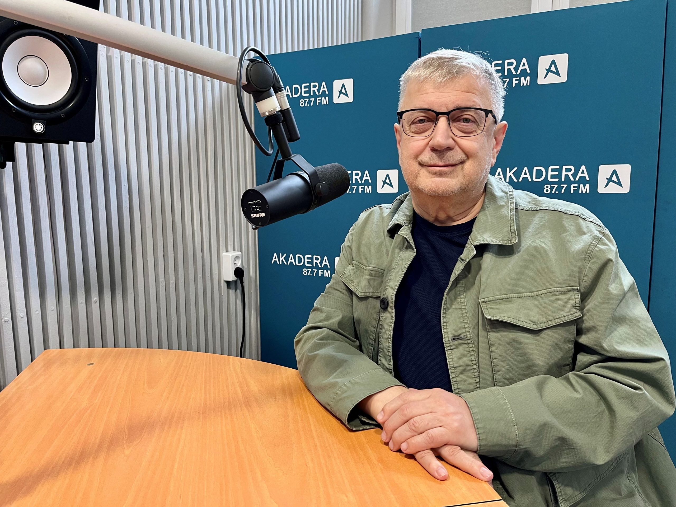 Prof. Joanicjusz Nazarko siedzi przy mikrofonie w studiu Radia Akadera. W tle niebieskie ścianki z logo „Akadera 87.7 FM”. Profesor ma na sobie oliwkową kurtkę i okulary, patrzy spokojnie w stronę obiektywu.
