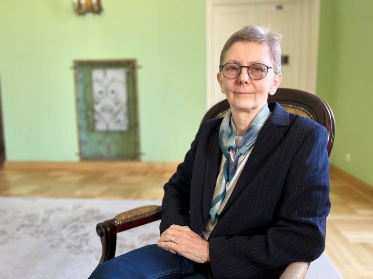 Prof. dr hab. n. med. Joanna Zajkowska siedzi na eleganckim, drewnianym fotelu w jasnym, stylowym pomieszczeniu. Ma na sobie ciemnogranatową marynarkę w delikatne prążki, jasnoniebieską apaszkę i okulary. Spogląda spokojnie w stronę obiektywu, z łagodnym wyrazem twarzy.