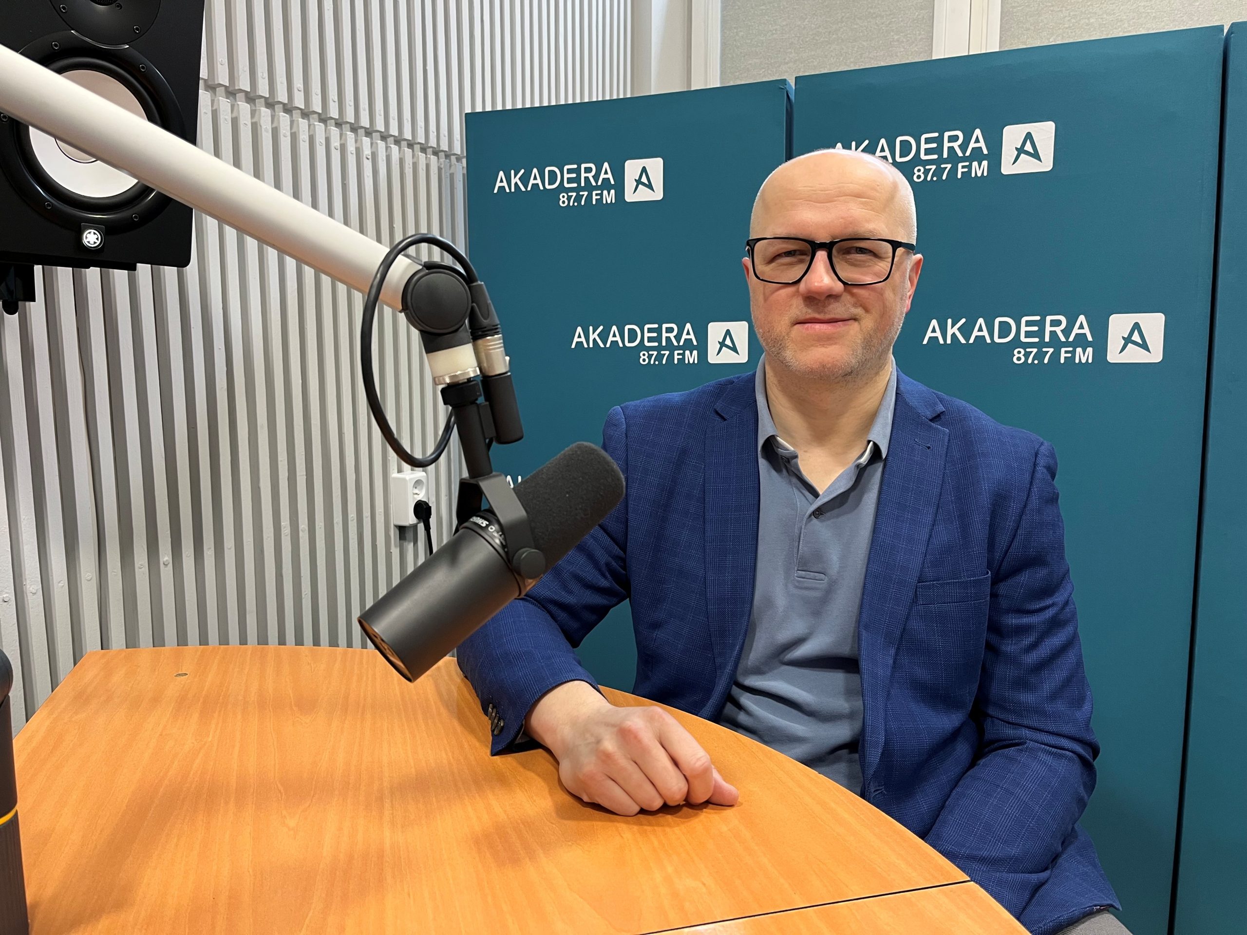 Dr inż. Paweł Tadejko siedzi przy mikrofonie w studiu Radia Akadera. W tle niebieskie ścianki z logo „AKADERA 87.7 FM”. Ma na sobie niebieską marynarkę i okulary.