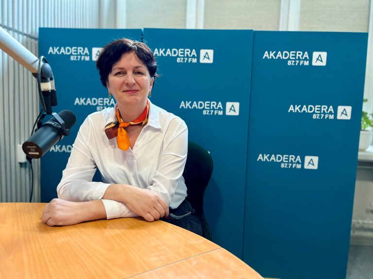 Prof. dr hab. inż. Agnieszka Dardzińska-Głębocka w studio Radia Akadera, siedzi przy mikrofonie, uśmiechnięta, w białej koszuli i kolorowej apaszce, na tle ścianki z logo stacji radiowej AKADERA 87.7 FM