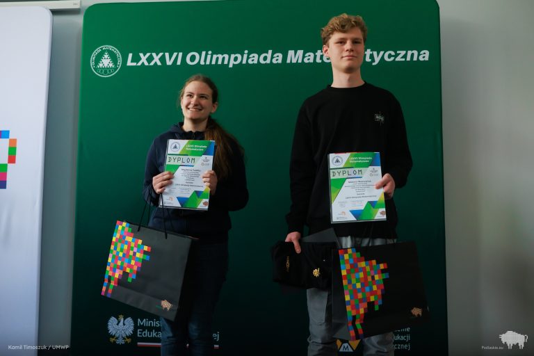 Dwoje młodych laureatów trzymających dyplomy zwycięzców Olimpiady Matematycznej. Magdalena Pudełko z uśmiechem, ubrana w ciemną bluzę, trzyma także torbę z kolorowym logo Podlaskiego. Obok niej stoi Mateusz Wawrzyniak z dyplomem i torbą w ręku. W tle zielona ścianka z napisem „LXXVI Olimpiada Matematyczna”.