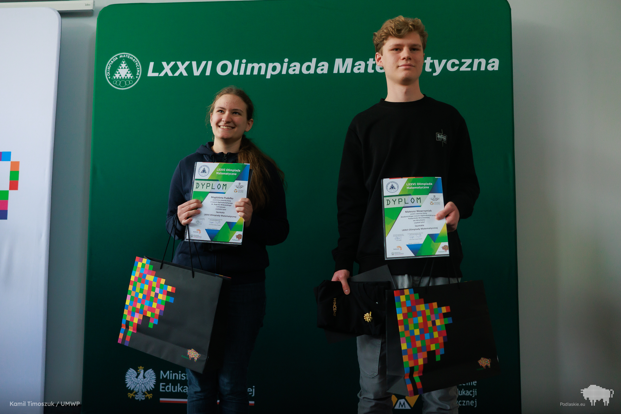 Dwoje młodych laureatów trzymających dyplomy zwycięzców Olimpiady Matematycznej. Magdalena Pudełko z uśmiechem, ubrana w ciemną bluzę, trzyma także torbę z kolorowym logo Podlaskiego. Obok niej stoi Mateusz Wawrzyniak z dyplomem i torbą w ręku. W tle zielona ścianka z napisem „LXXVI Olimpiada Matematyczna”.