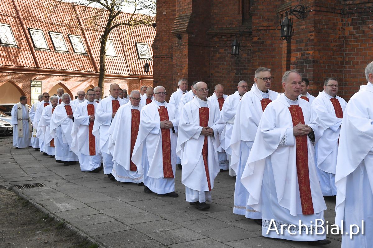 Grupa kilkudziesięciu księży ubranych w białe alby z czerwonymi stułami uczestniczy w uroczystej procesji przed katedrą w Białymstoku z okazji Wielkiego Czwartku. W tle widoczne ceglane mury świątyni i budynki Arkad Katedralnych.