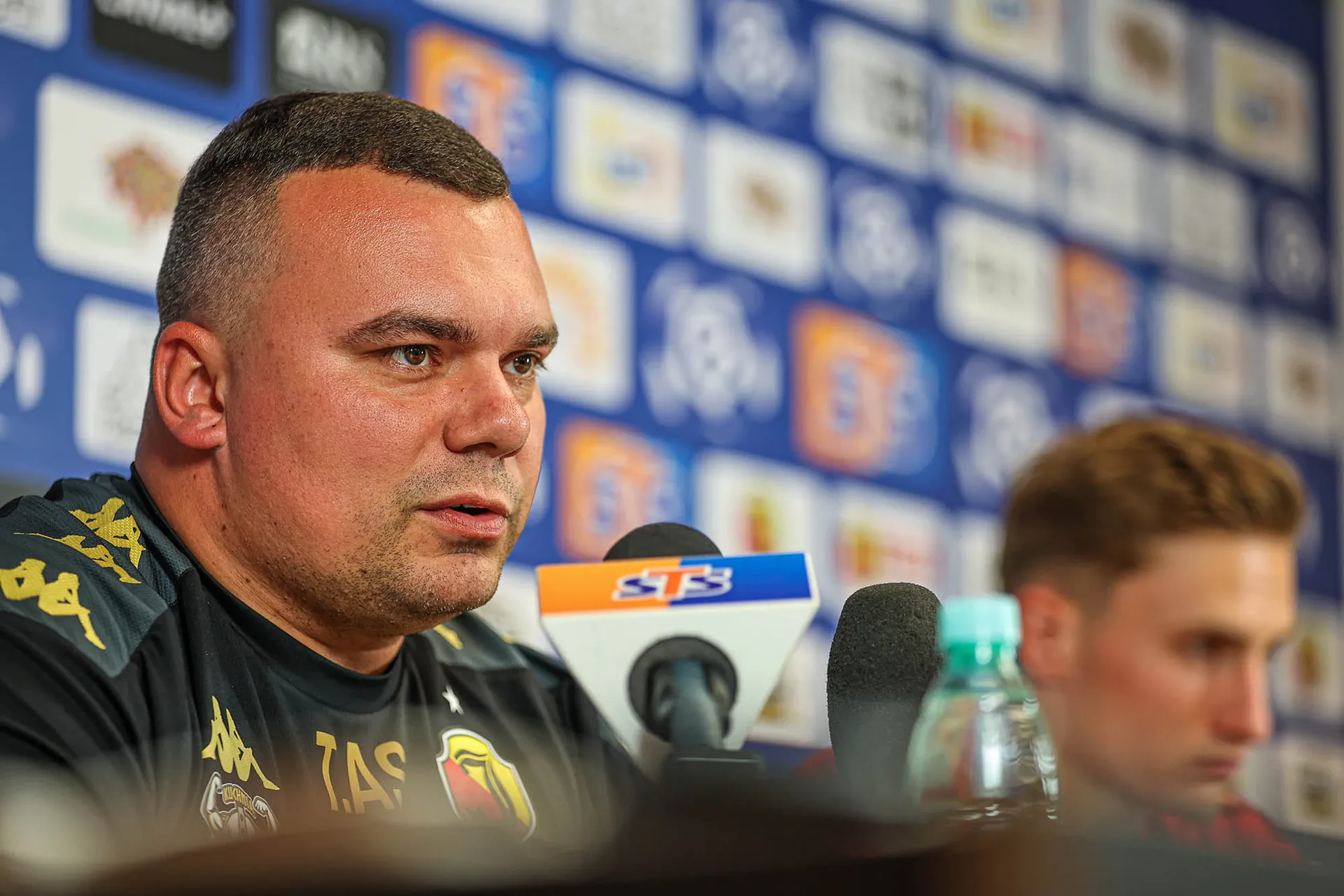 Adrian Siemieniec, trener Jagiellonii Białystok, w czarnej koszulce z herbem klubu i logotypami sponsorów, mówi do mikrofonów podczas konferencji prasowej. W tle ścianka z logotypami medialnymi i sponsorskimi. Obok niego rozmyta sylwetka piłkarza.
