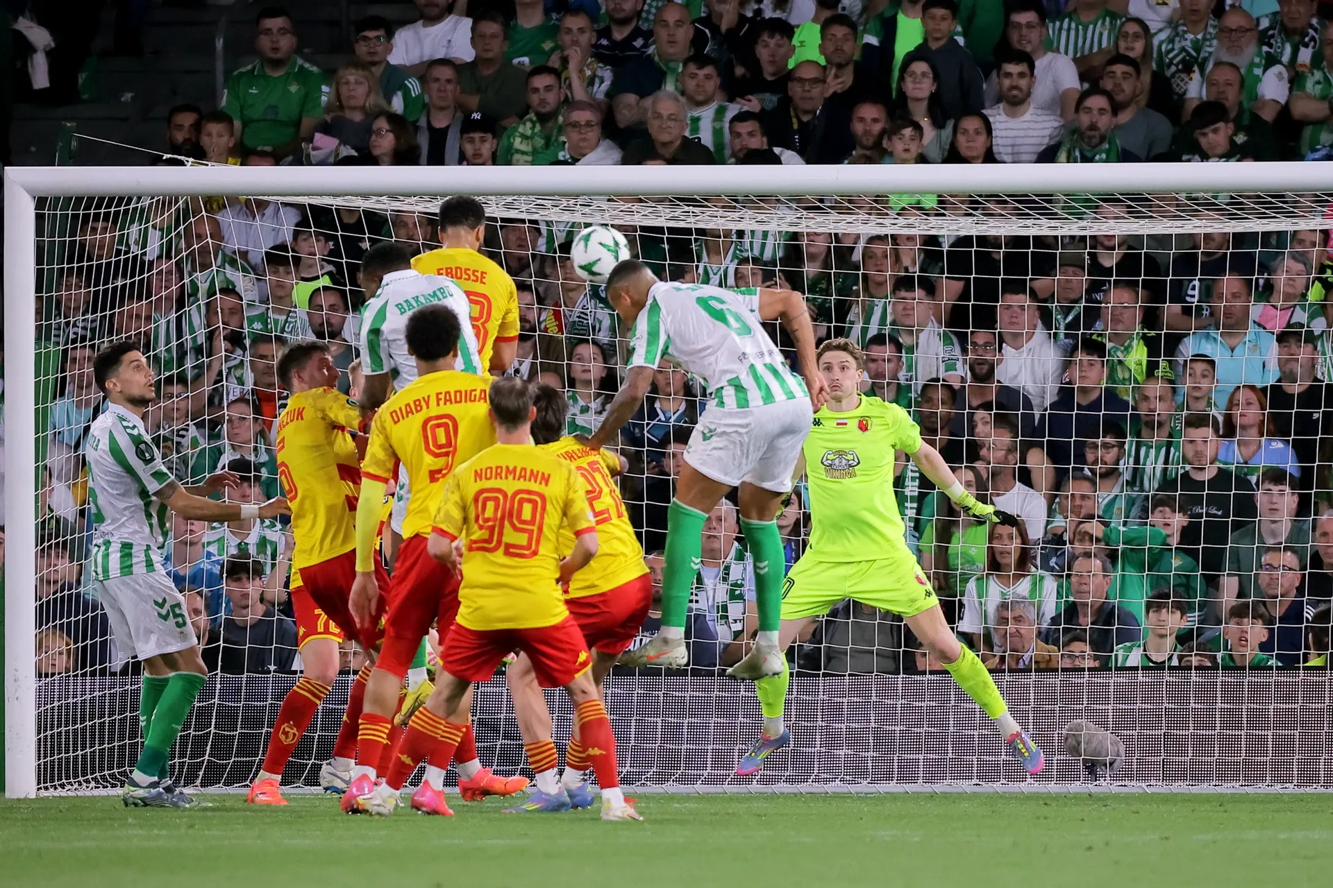 Piłkarze Realu Betis i Jagiellonii Białystok w akcji podbramkowej – zawodnik Betisu wyskakuje do główki, bramkarz Jagiellonii przygotowany do interwencji. W tle tłum kibiców w zielono-białych barwach zasiadających na trybunach stadionu Benito Villamarín w Sewilli.