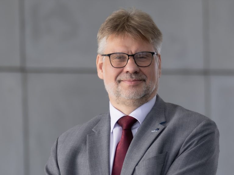 Zdjęcie przedstawia uśmiechniętego mężczyznę w okularach, w garniturze z czerwonym krawatem. Tło jest jednolite, szare. Na zdjęciu znajduje się dr hab. inż. Andrzej Gajewski.