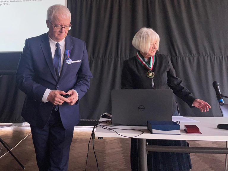 Profesor Jerzy Uścinowicz w garniturze z odznaczeniem na piersi stoi obok kobiety z siwymi włosami uhonorowanej medalem na wstędze. Oboje znajdują się przy stole z laptopem i mikrofonem, na tle czarnej kotary.