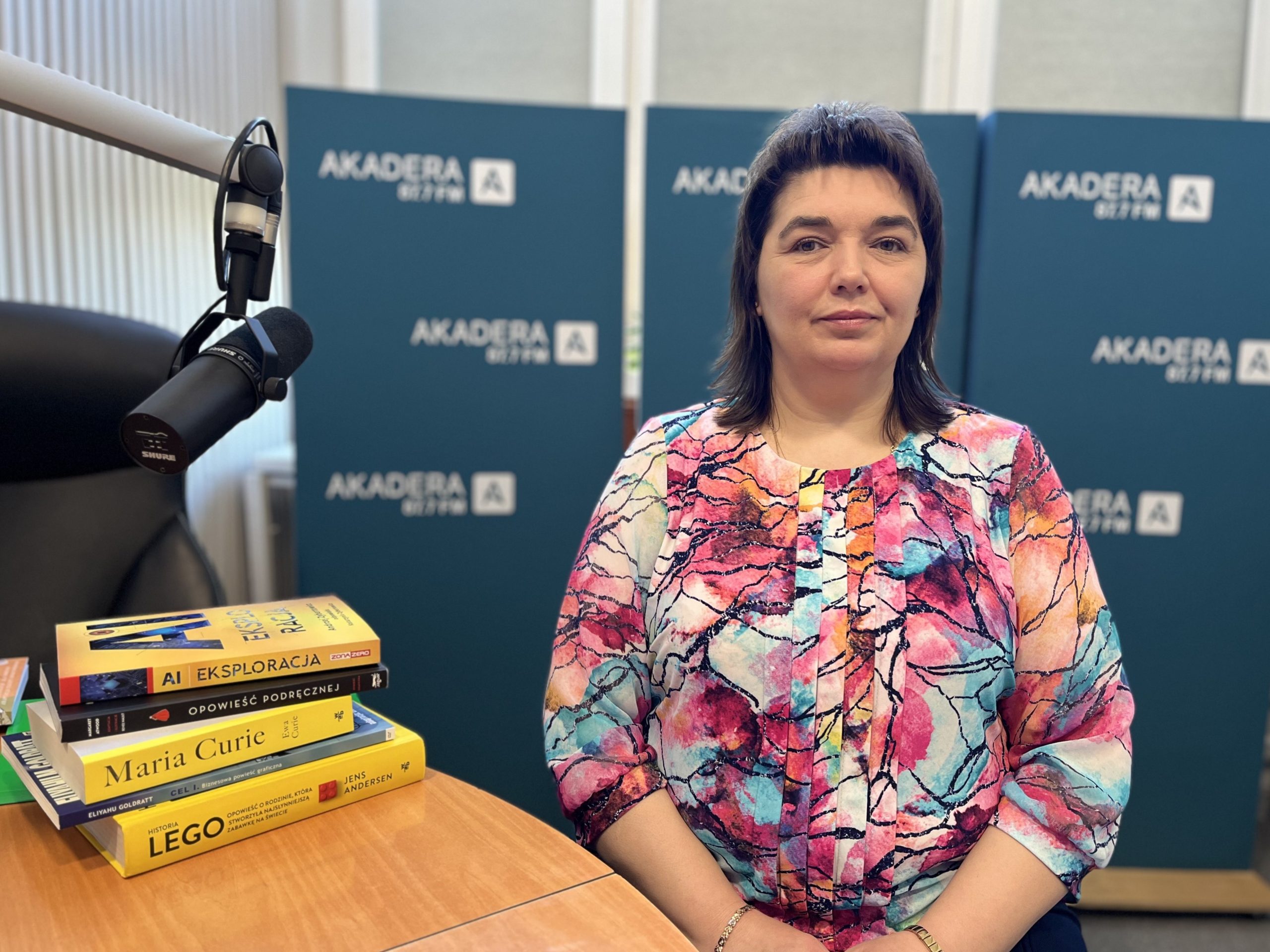 Kobieta w kolorowej bluzce siedzi przy mikrofonie radiowym w studiu Radia Akadera. Obok niej leży stos książek. W tle plansze z logo Radia Akadera.