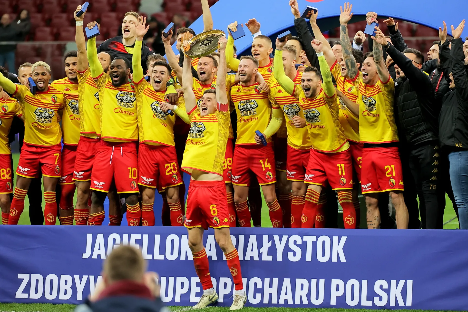 Piłkarze Jagiellonii Białystok w żółto-czerwonych strojach świętują zdobycie Superpucharu Polski. Na środku zawodnik z numerem 6 unosi trofeum w górę, wokół niego zespół wiwatuje z uniesionymi rękami. W tle widoczne są trybuny Stadionu Narodowego.