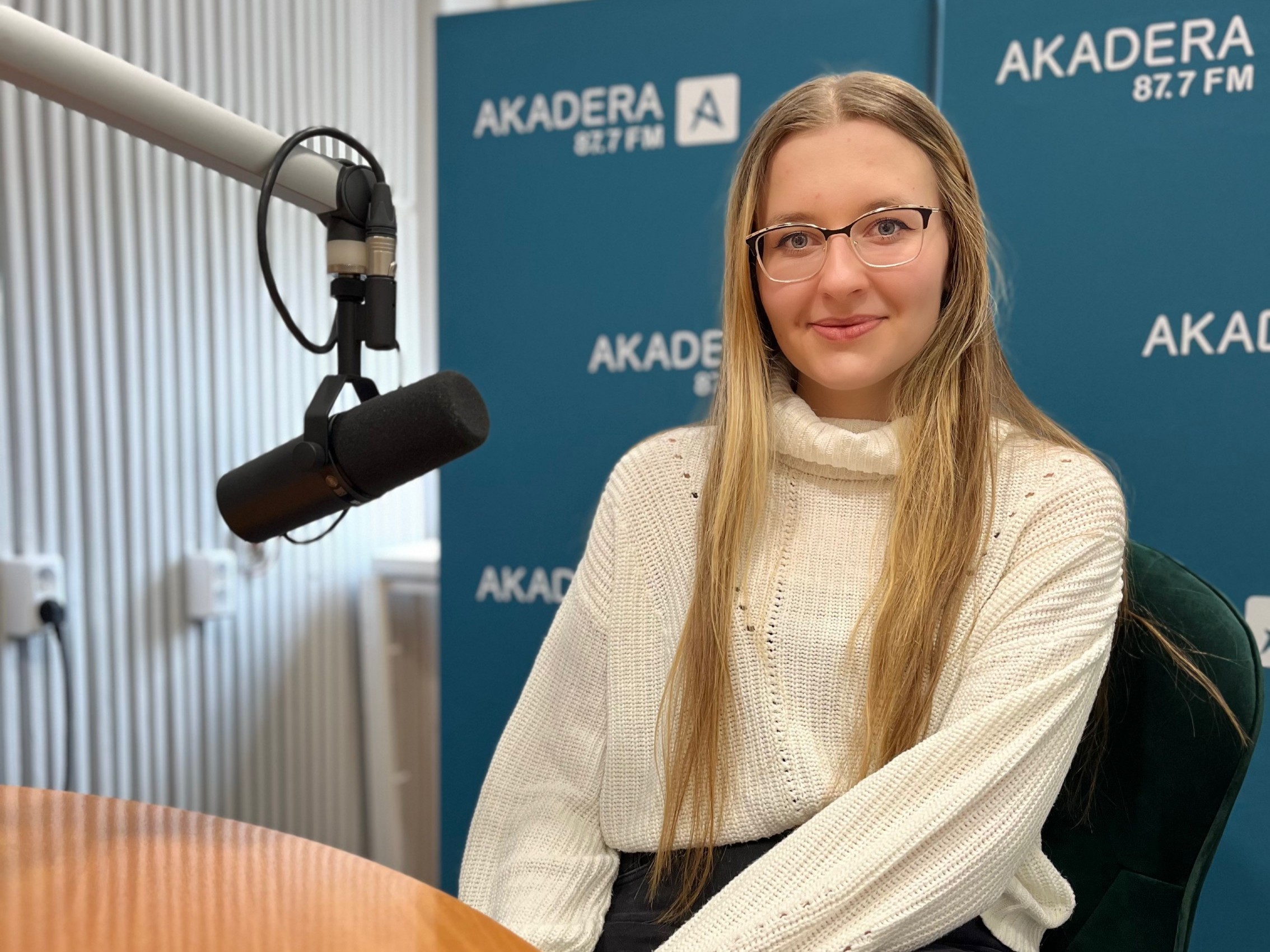 Młoda kobieta z długimi blond włosami i okularami, ubrana w jasny sweter, siedzi przy stole radiowym w studiu Radia AKADERA. Obok niej mikrofon studyjny. W tle ścianka z logo AKADERA 87.7 FM.