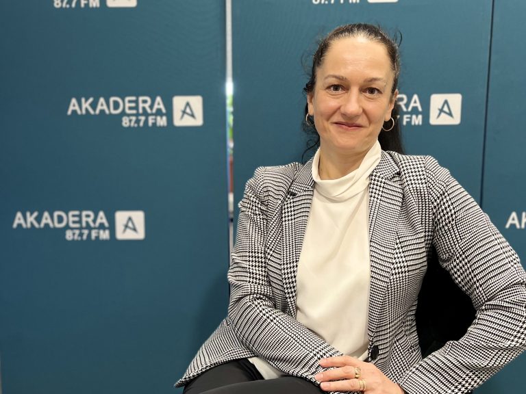 Prof. Katarzyna Czerewacz-Filipowicz siedzi w studiu Radia Akadera, ubrana w biały golf i elegancką kraciastą marynarkę. W tle widoczne są niebieskie ścianki z logo Akadera 87.7 FM.
