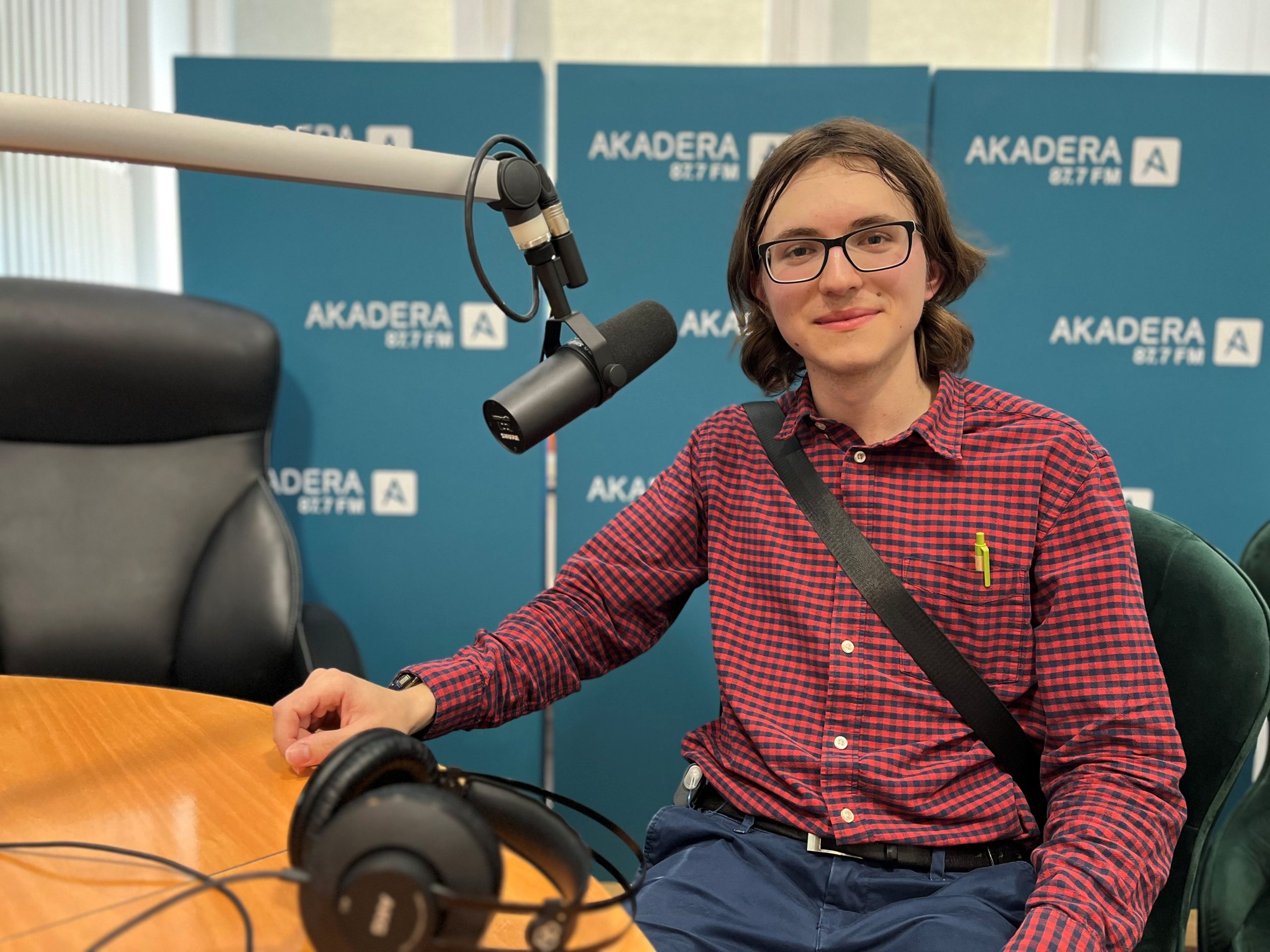 Młody mężczyzna w okularach i czerwonej koszuli w kratę siedzi przy stole radiowym w studiu Radia AKADERA. Przed nim mikrofon i słuchawki, w tle ścianka z logo AKADERA 87.7 FM.