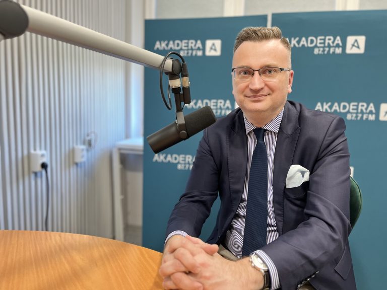 Rafał Rudnicki, zastępca prezydenta Białegostoku, siedzi w studiu Radia Akadera przy mikrofonie. Ubrany w garnitur, koszulę w paski i granatowy krawat, patrzy w stronę obiektywu. W tle widoczna ścianka z logo Akadera 87,7 FM.