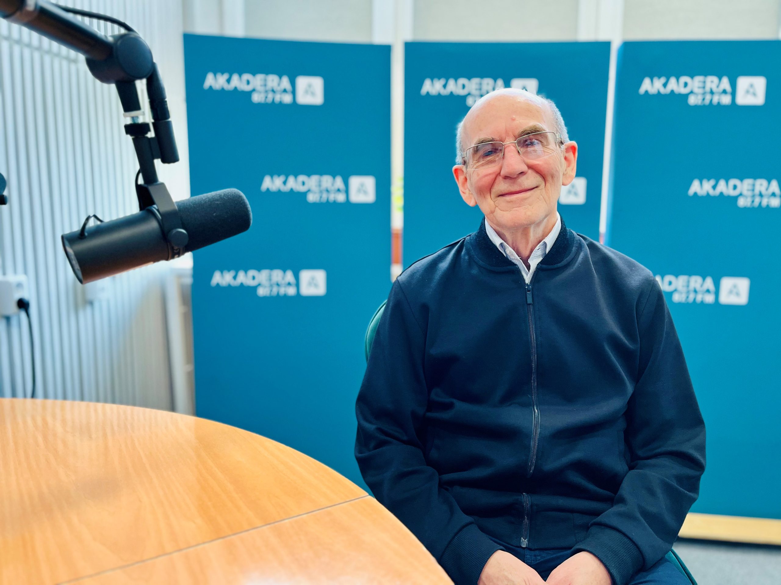 Profesor Zbigniew Bartosiewicz siedzi w studiu Radia AKADERA. W tle widoczne niebieskie ścianki z logo AKADERA 87.7 FM. Po lewej stronie widoczny mikrofon studyjny. Profesor ma na sobie granatową bluzę i uśmiecha się do kamery.