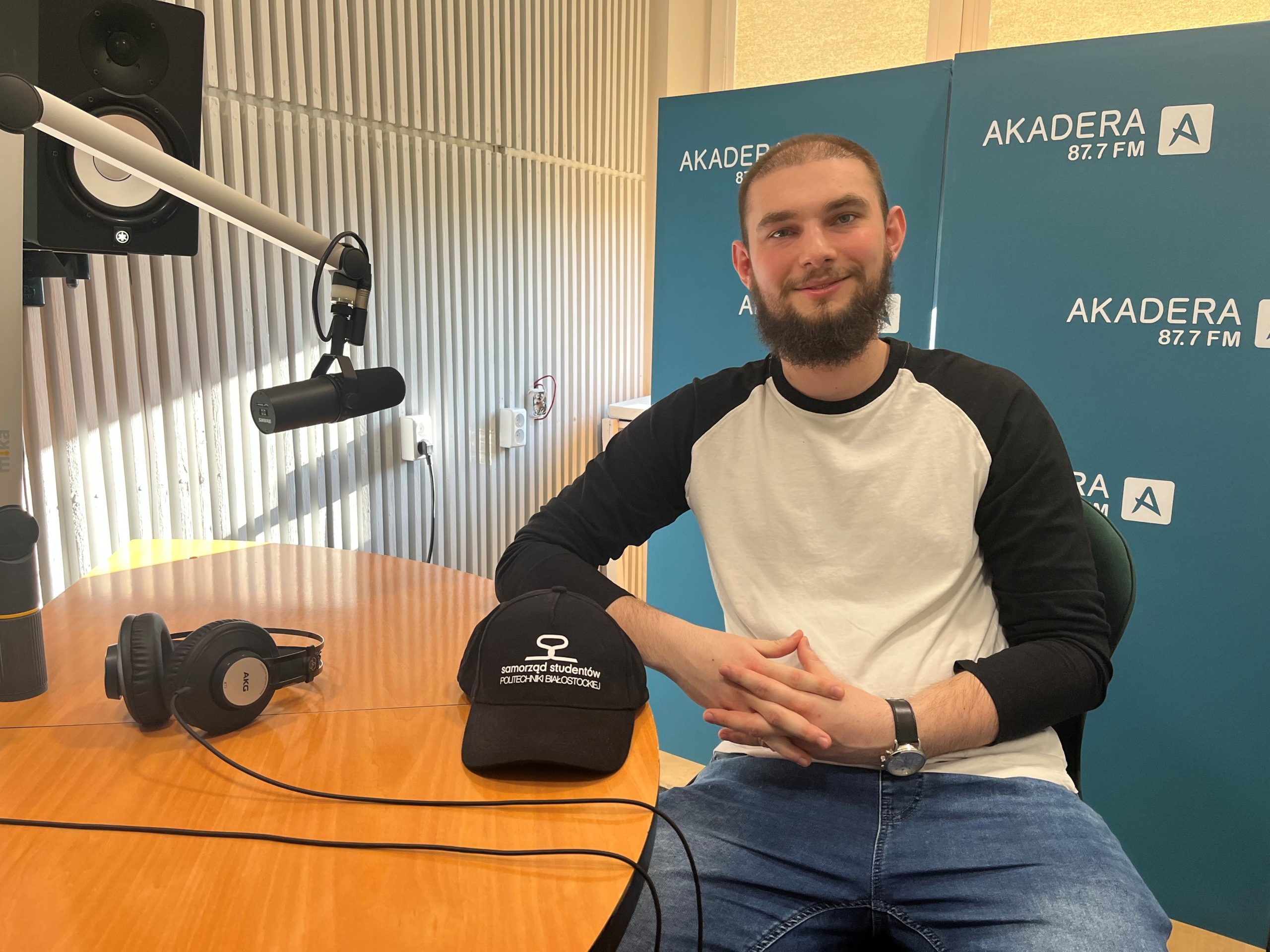 Uśmiechnięty młody mężczyzna z brodą, ubrany w biało-czarną koszulkę z długim rękawem, siedzi w studiu radiowym Radia AKADERA. Przed nim na stole leży czapka z logo samorządu studentów Politechniki Białostockiej oraz słuchawki. W tle widoczne są panele dźwiękochłonne i banery z logo AKADERA 87.7 FM.