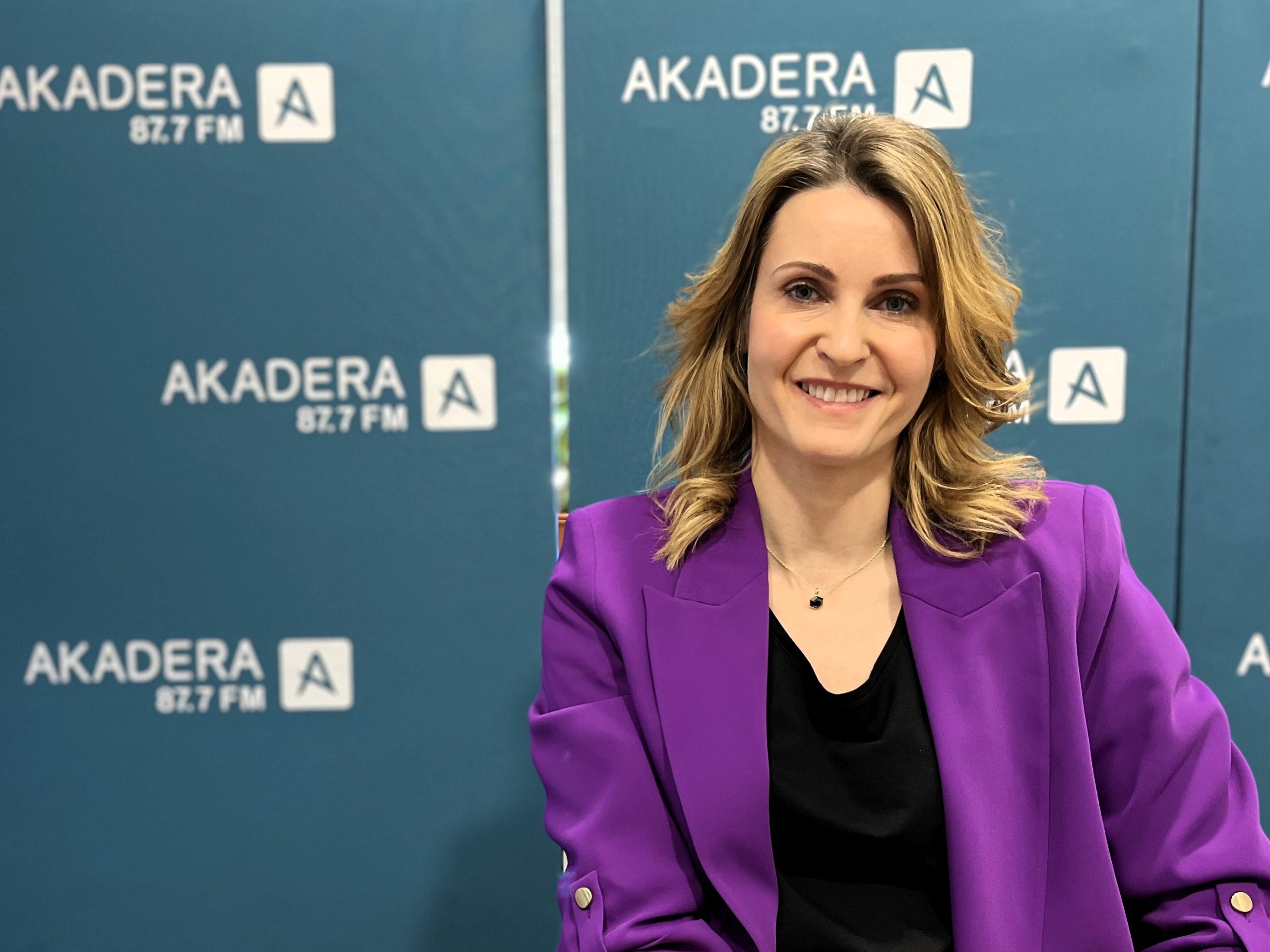 Dr hab. Monika Kalinowska w studiu radiowym Radia AKADERA, ubrana w fioletową marynarkę i czarną bluzkę. W tle widoczne są zielone ścianki z logo Radia AKADERA 87.7 FM. Dr Kalinowska uśmiecha się i patrzy prosto w obiektyw.