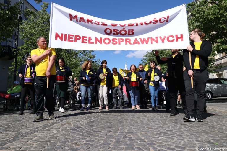 Duży czerwony transparent z białym napisem „MARSZ OSÓB NIEPEŁNOSPRAWNYCH” niesiony przez uczestników. W tle flaga Polski i kamienice.