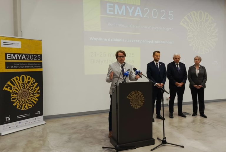 Zdjecie z konferencji zapowiadajcej Kongres Muzealnikow, na srodku stoi 5 osob, mezczyzna przemawia, pozostali stoja obok, w tle logo EMYA