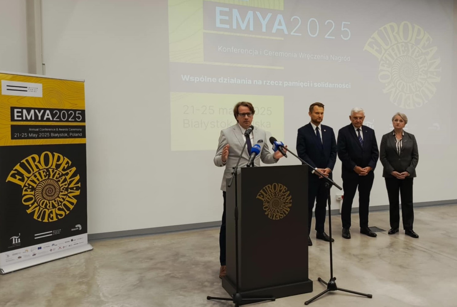 Zdjecie z konferencji zapowiadajcej Kongres Muzealnikow, na srodku stoi 5 osob, mezczyzna przemawia, pozostali stoja obok, w tle logo EMYA