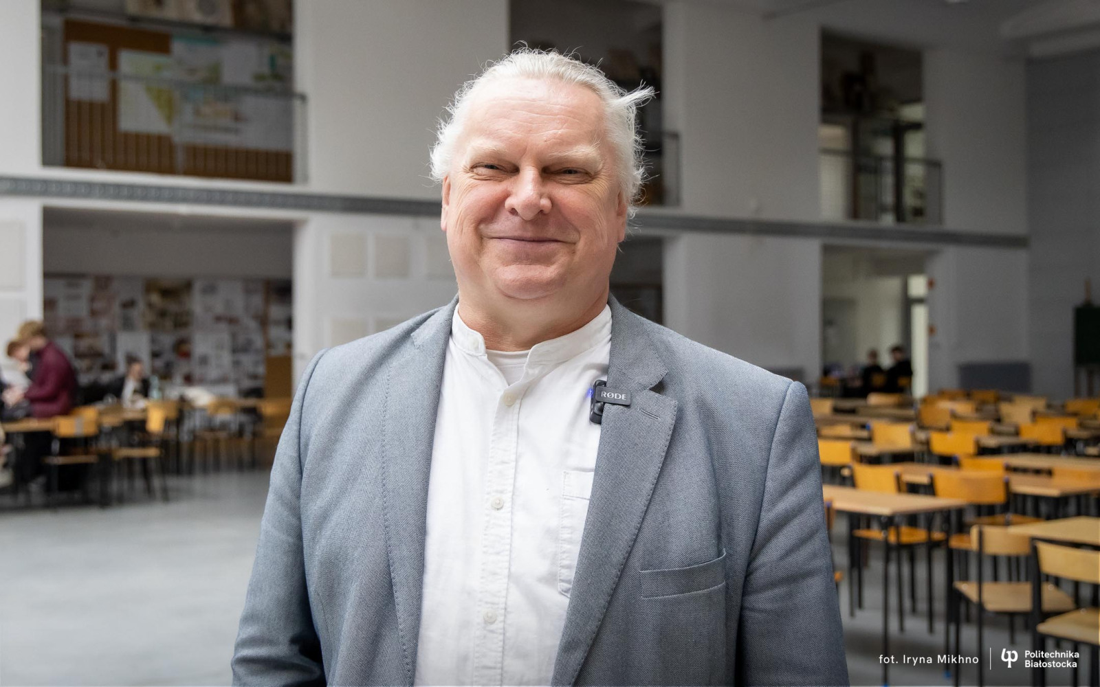 Dr inż. arch. Adam Jakimowicz, ubrany w szarą marynarkę i białą koszulę, uśmiecha się do obiektywu. W tle widać przestronną salę z ustawionymi rzędami drewnianych krzeseł i stołów oraz studentów pracujących nad projektami.