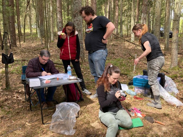 Zespół Magna Silva przy stole i w terenie dokumentuje znalezisko monet w lesie – archeologiczna dokumentacja w Puszczy Knyszyńskiej.