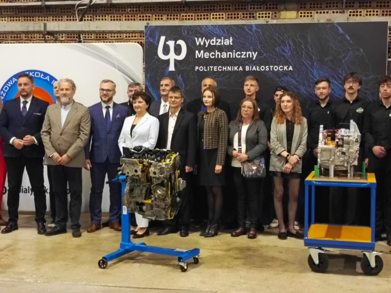 Na tle ścianki z napisem wydzial mechaniczny politechniki bialostockiej stoja elegancko ubrani przedstawciele swiata nauki i biznesu. Przed nimi dwa przekroje silnikow, majacych sluzyc jako pomoze dydaktyczne
