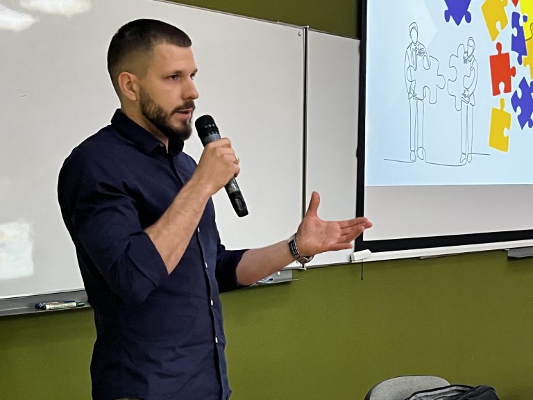 Mężczyzna z mikrofonem w ręku przemawia do studentów na tle prezentacji z rysunkiem puzzli i sylwetek ludzi.