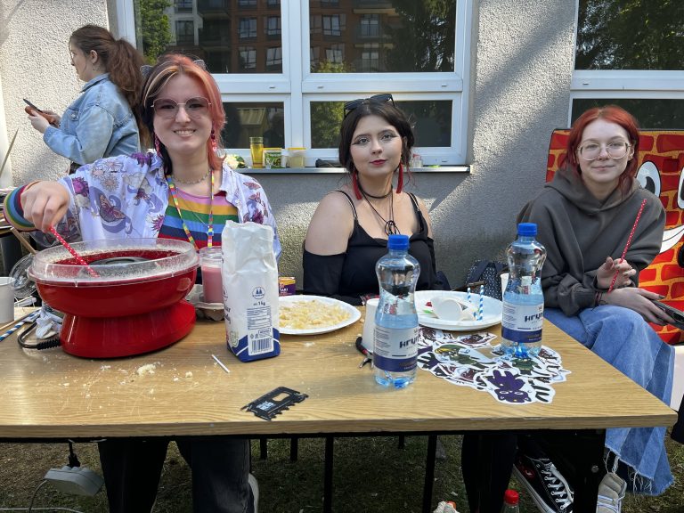 Trzy studentki Wydziału Architektury PB siedzące przy stole z watą cukrową i przekąskami, uśmiechnięte i ubrane kolorowo.