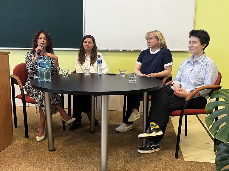 Cztery kobiety – uczestniczki panelu dyskusyjnego – siedzą przy okrągłym stole podczas debaty o równości i inkluzywności. Jedna z nich mówi do mikrofonu. W tle tablica i jasnożółta ściana sali wykładowej Politechniki Białostockiej.