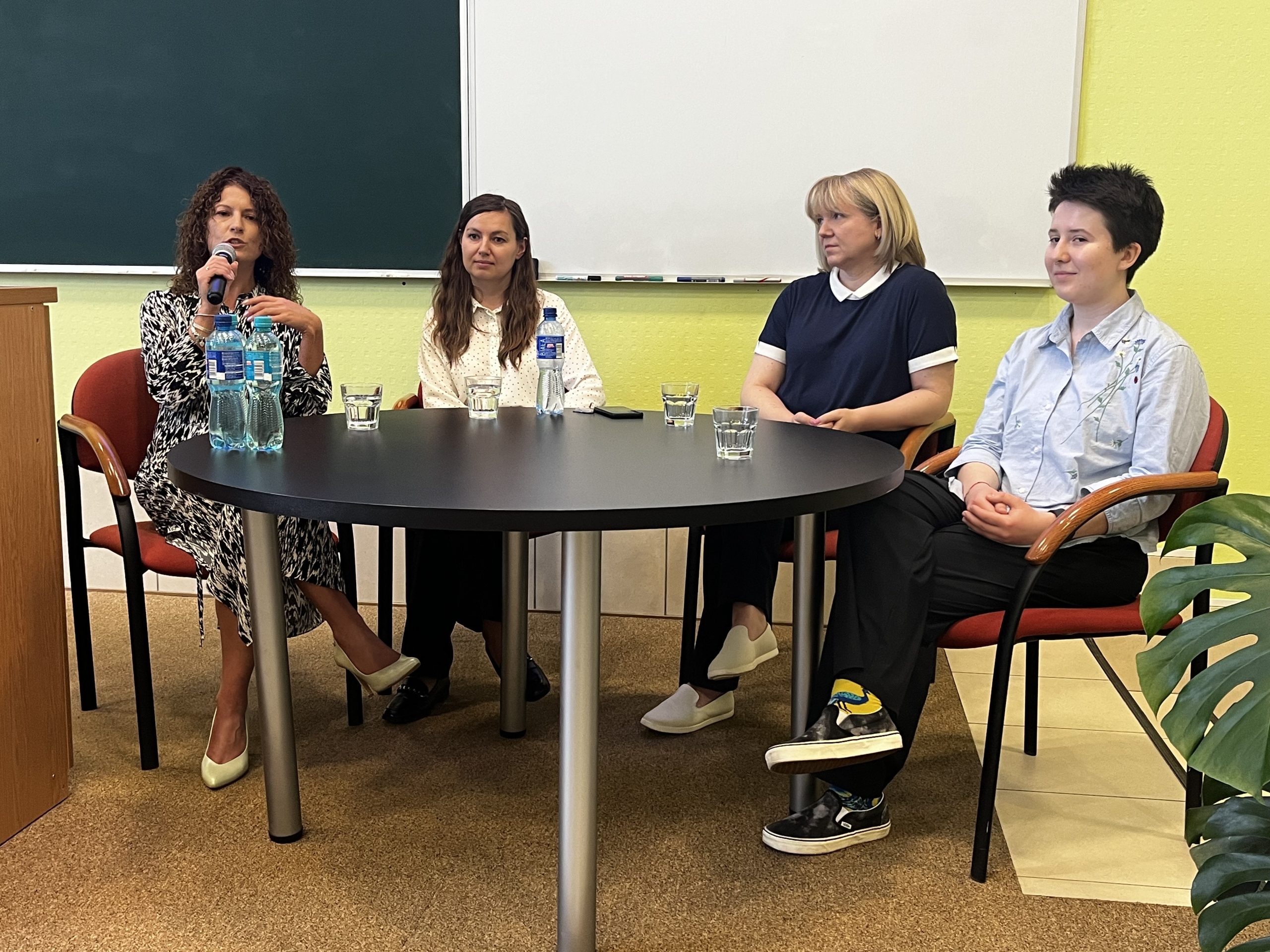 Cztery kobiety – uczestniczki panelu dyskusyjnego – siedzą przy okrągłym stole podczas debaty o równości i inkluzywności. Jedna z nich mówi do mikrofonu. W tle tablica i jasnożółta ściana sali wykładowej Politechniki Białostockiej.