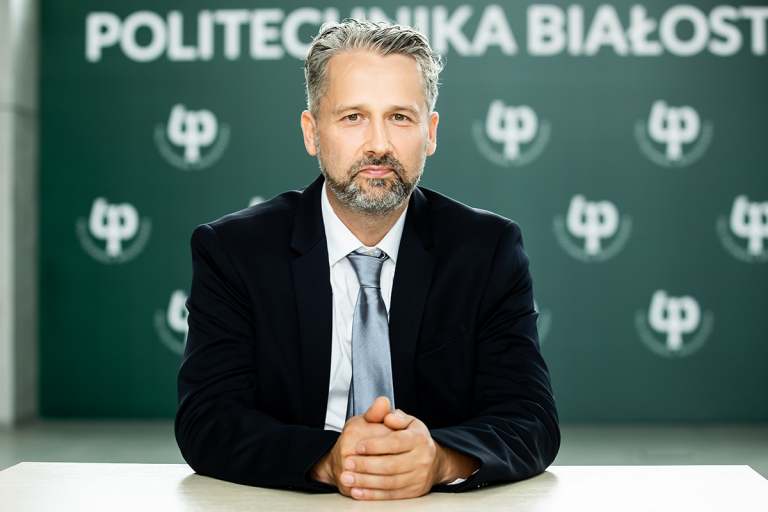 Dr hab. inż. Jarosław Szusta w garniturze, siedzący przy stole na tle logotypu Politechniki Białostockiej, patrzy w obiektyw.